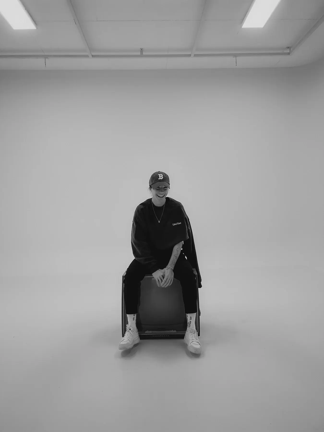 Corinna Kasus seated in a bright room, dressed in black, wearing a cap.